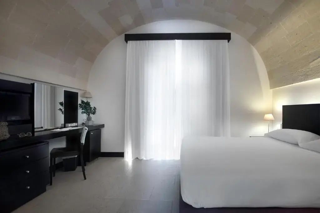 Hotelzimmer mit weißem Gewölbe, langen Gardinen an den Fenstern und Schreibtisch.