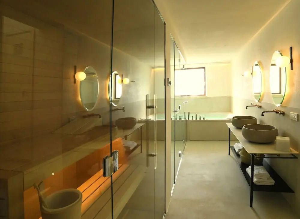 Spa Bereich mit Glasabtrennung, Holzsauna, Doppelwaschtisch und Whirlpool Wanne im Hintergrund.