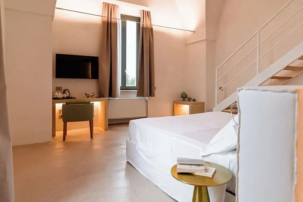 Doppelbett mit weißer Bettwäsche, Beistelltisch und Arbeitsbereich im hellen Hotelzimmer.