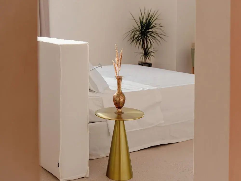 Helles Hotelzimmer mit weiß bezogenem Bett, goldenem Beistelltisch und einer Zimmerpflanze.
