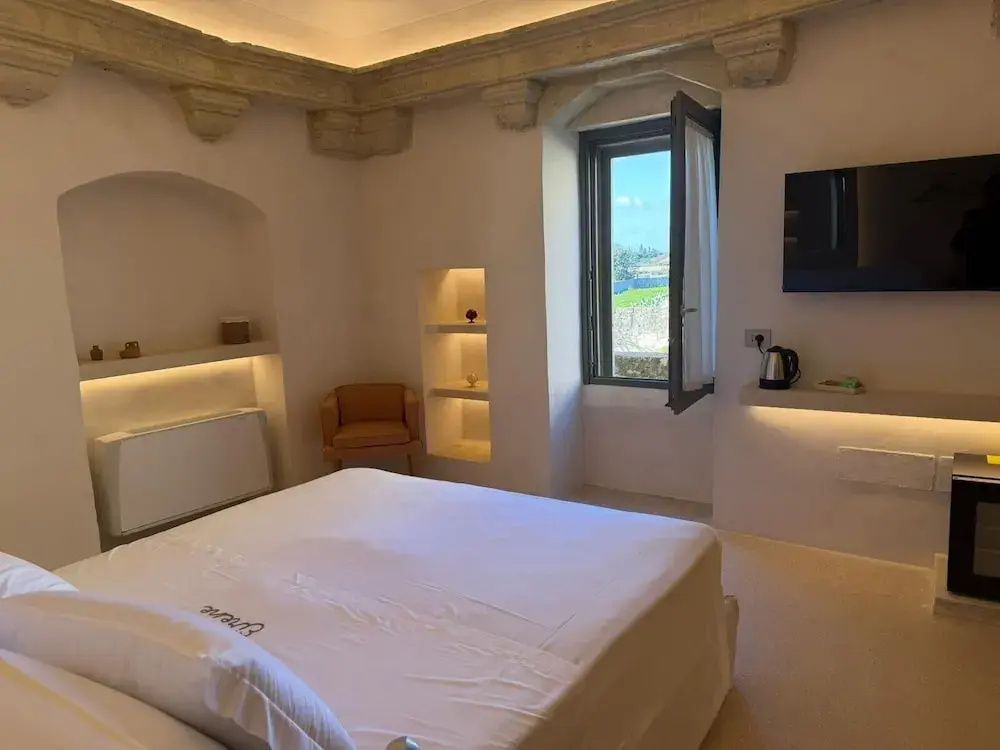 Schlafzimmer mit weiß eingedecktem Bett, Sessel, beleuchteten Nischen und Fenster.