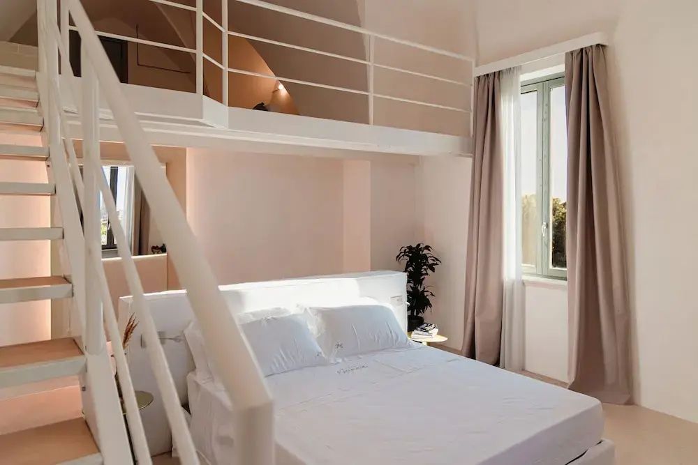 Helles Hotelzimmer mit weißem Doppelbett, Vorhängen am Fenster und einer Empore.