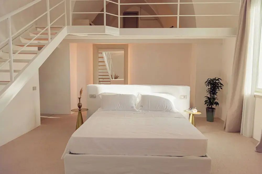 Weißes Bett unter einem gewölbten Raum mit Empore, Treppe und Beistelltischen.