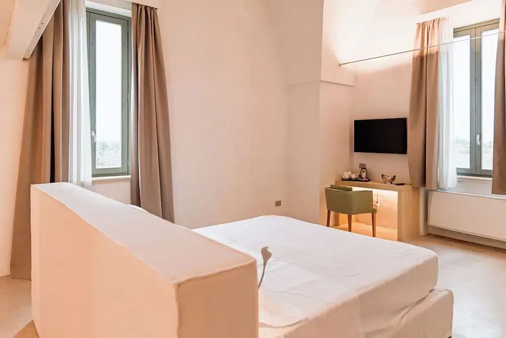 Doppelbett in hellem Hotelzimmer mit Schreibtisch, grünem Stuhl und Wandfernseher.