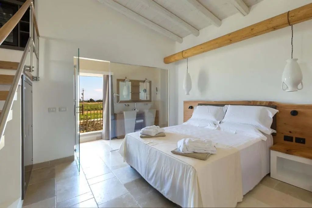 Helles Schlafzimmer mit weißem Doppelbett, Glasabtrennung zum Bad und Blick ins Freie.