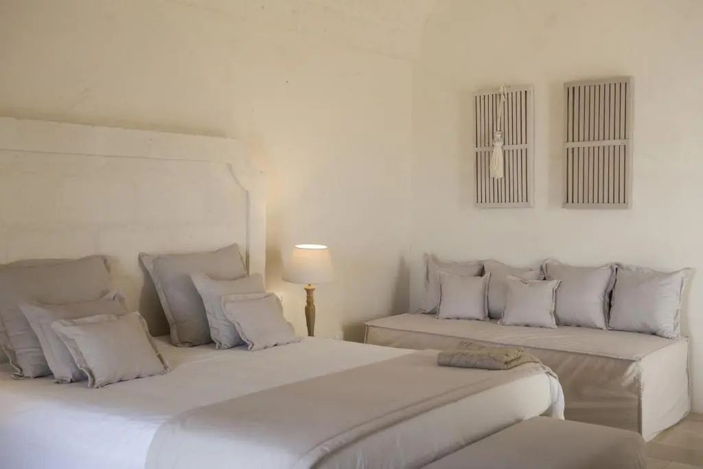 Schlafbereich mit weißer Wand, großem Bett, Nachttischlampe und einer weiteren Sitzgelegenheit.