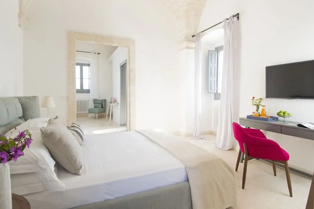 Weiß eingerichtetes Hotelzimmer mit Doppelbett, einem pinken Stuhl am Schreibtisch und Durchgang.