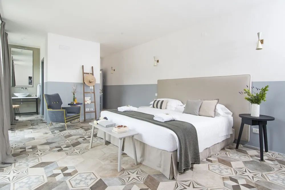 Großes Doppelbett mit grauer Tagesdecke, Holzbank davor und kunstvollem Fliesenboden im Hotelzimmer.