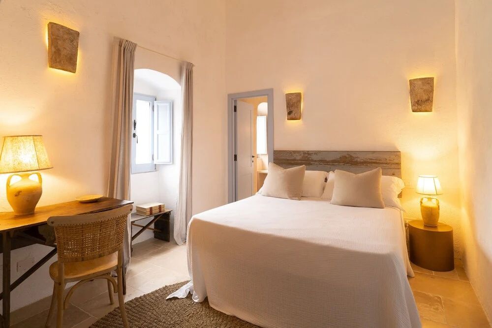 Schlafzimmer mit einem Doppelbett, kleinem Schreibtisch und Bogenfenster im Hintergrund.
