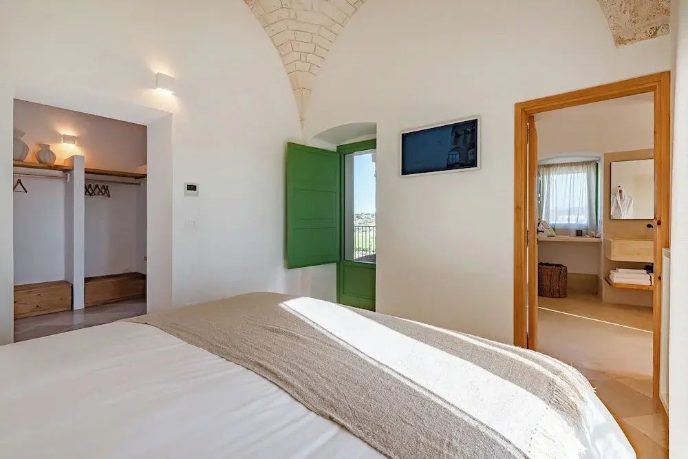 Hell eingerichtetes Schlafzimmer mit Gewölbedecke, Doppelbett, Fernseher und grüner Tür nach draußen.