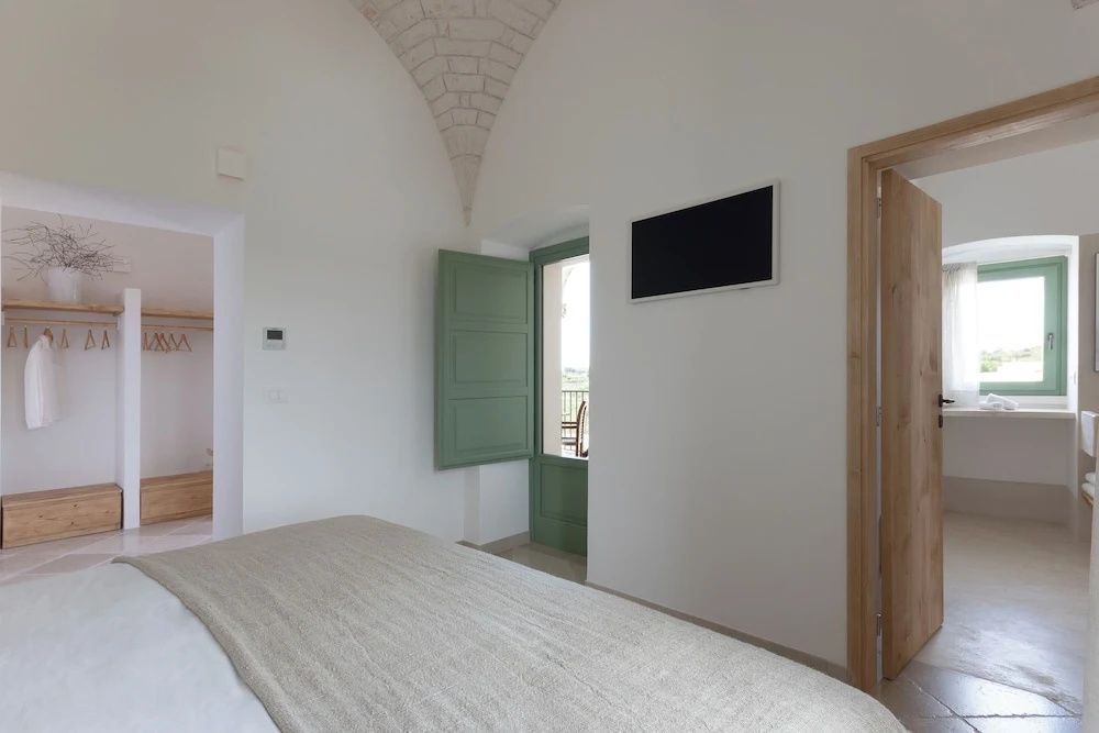 Hell gestaltetes Hotelzimmer mit Doppelbett, TV an der Wand und Zugang zum Balkon mit grüner Tür.