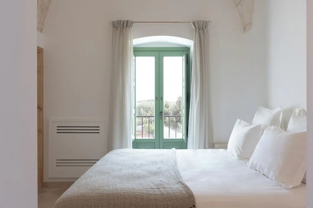 Helles Doppelbett mit weißer Bettwäsche vor grünem Fenster mit hellen Vorhängen.