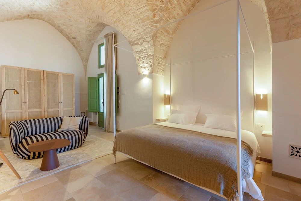 Hotelzimmer mit weißem Himmelbett, gestreiftem Sofa, Steingewölbe und grünen Fensterläden.