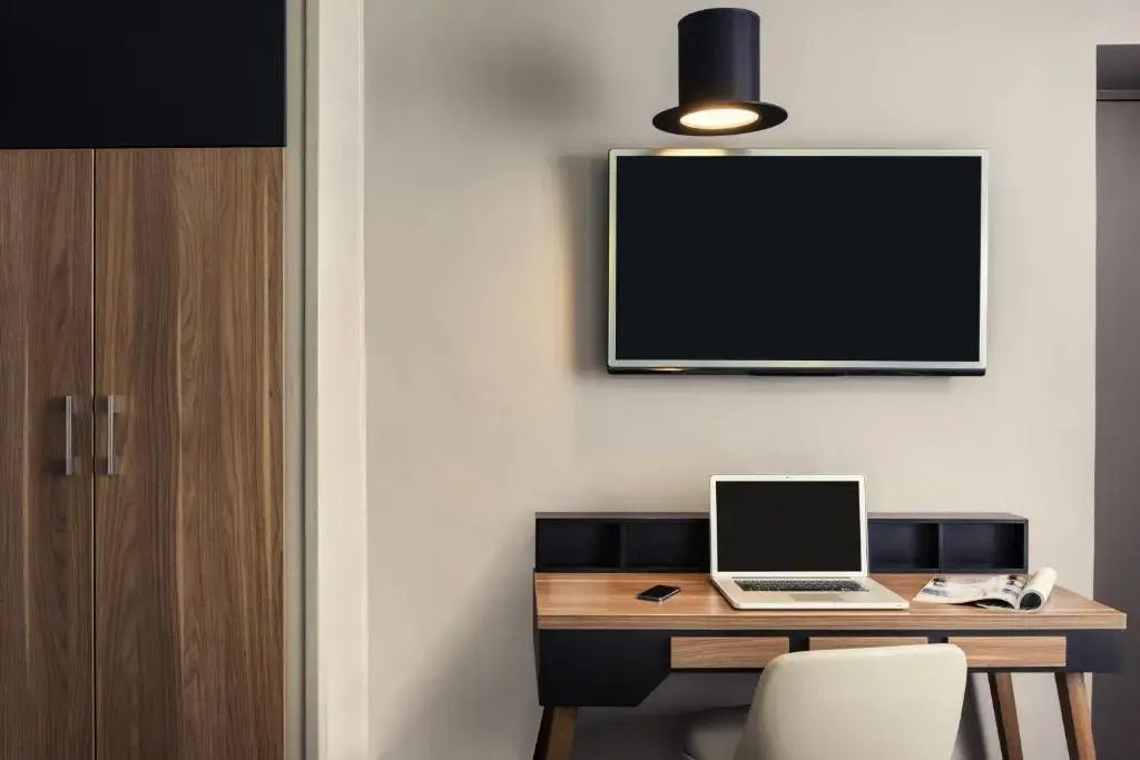 Schreibtisch aus Holz mit Laptop, Wandfernseher und einer Deckenlampe in Zylinderform.