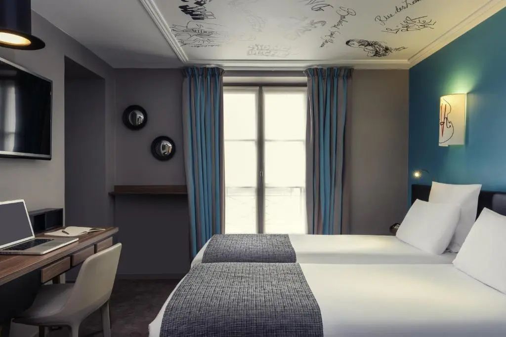 Blick in ein Hotelzimmer mit zwei Einzelbetten, Schreibtisch und großem Fenster mit Vorhängen.