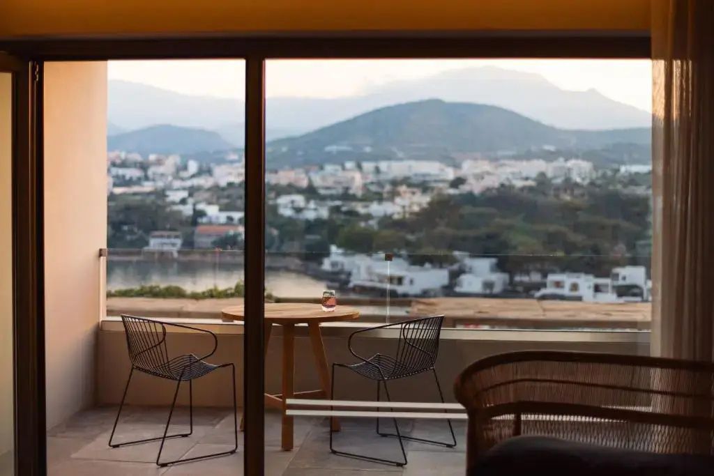 Sicht durch ein Fenster auf einen Balkon mit Sitzgelegenheiten und eine Küstenstadt.
