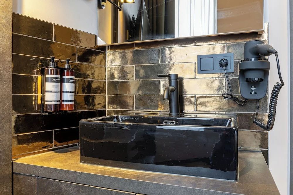 Schwarzes eckiges Waschbecken mit schwarzer Armatur vor dunklen glänzenden Fliesen.