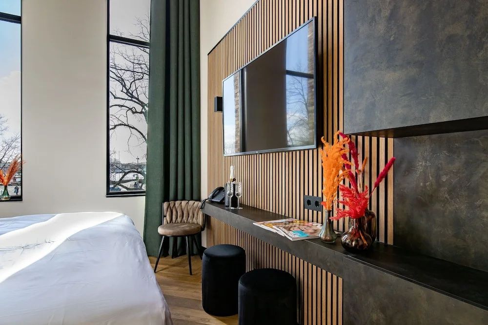 Flachbildfernseher an einer Holzlamellenwand mit Ablage, Hockern und Stuhl im Hotelzimmer.