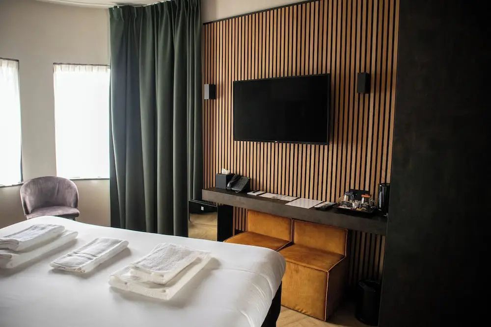 Wandfernseher an einer Holzlamellenwand mit Schreibtisch und gelben Sitzhockern.