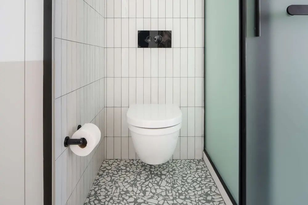 Weiß gefliestes Badezimmer mit einer wandhängenden Toilette und hellgrauem Mosaikfußboden.