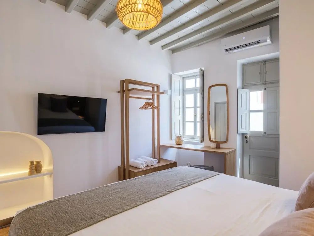 Helles Schlafzimmer mit Doppelbett, offenem Kleiderständer aus Holz, Fernseher und Spiegel.