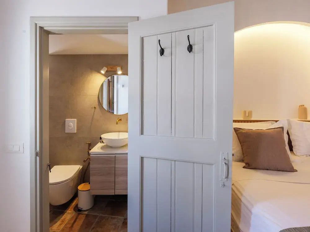 Blick durch eine weiße Holztür auf ein modernes Badezimmer mit rundem Spiegel und Waschtisch.
