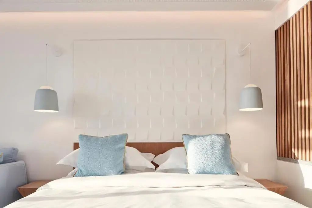 Doppelbett mit weißen Kissen und hellblauen Akzentkissen vor weiß strukturierter Wand.