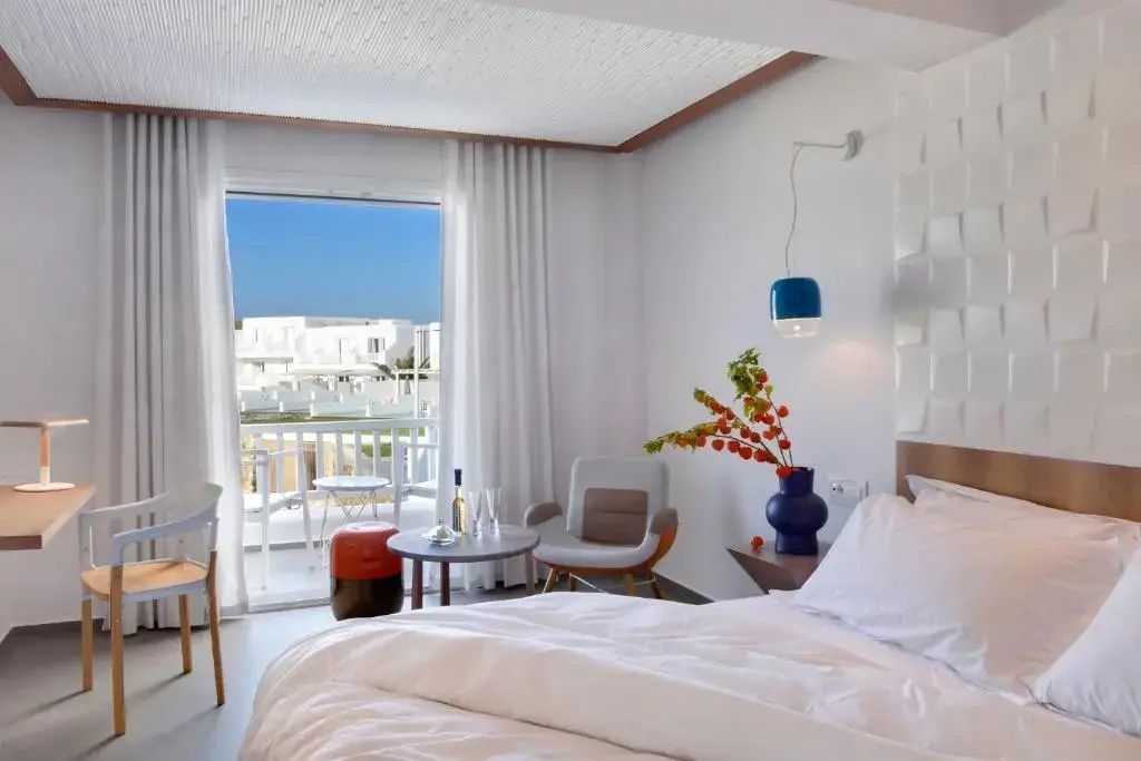 Helles Hotelzimmer mit Doppelbett, Sessel, Deko-Vase und Zugang zum Balkon.