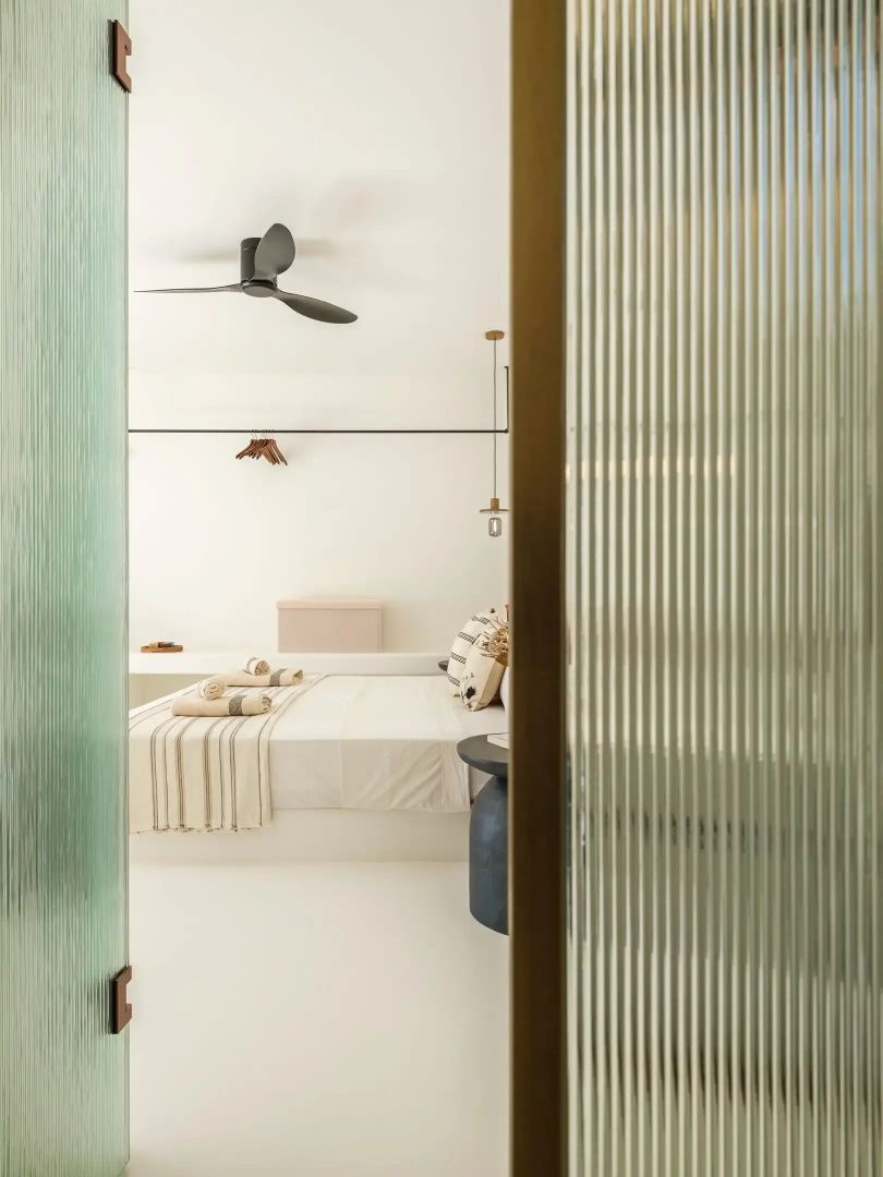 Blick durch eine geriffelte Glastür in ein helles Schlafzimmer mit Doppelbett und Deckenventilator.