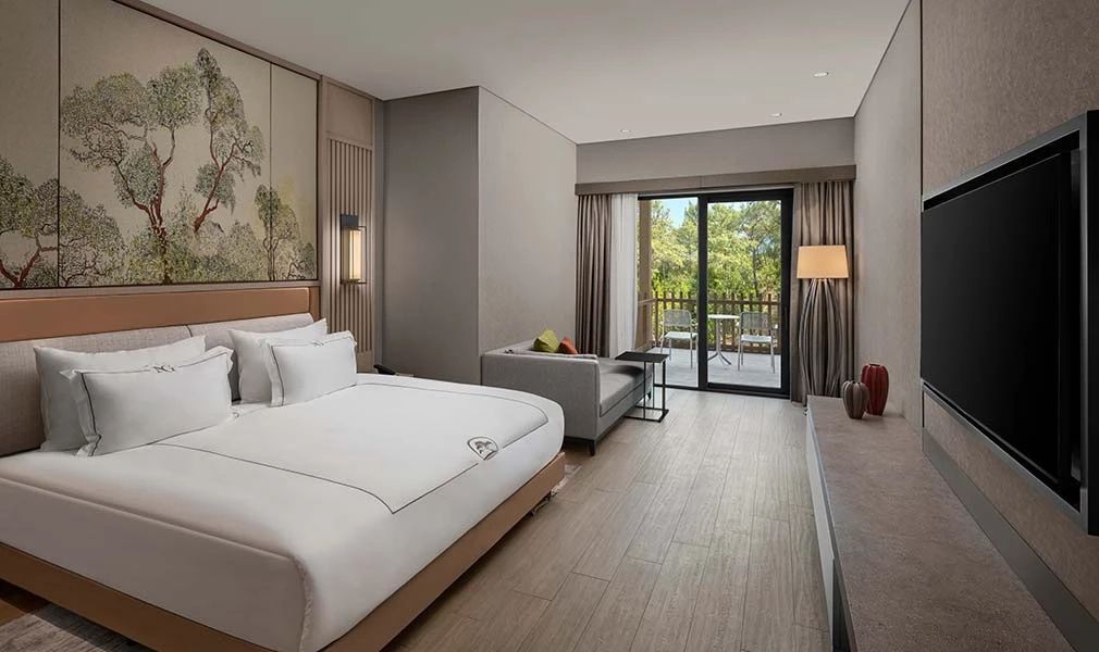 Hotelzimmer mit Doppelbett, grauem Sofa und Balkonzugang ins Grüne.