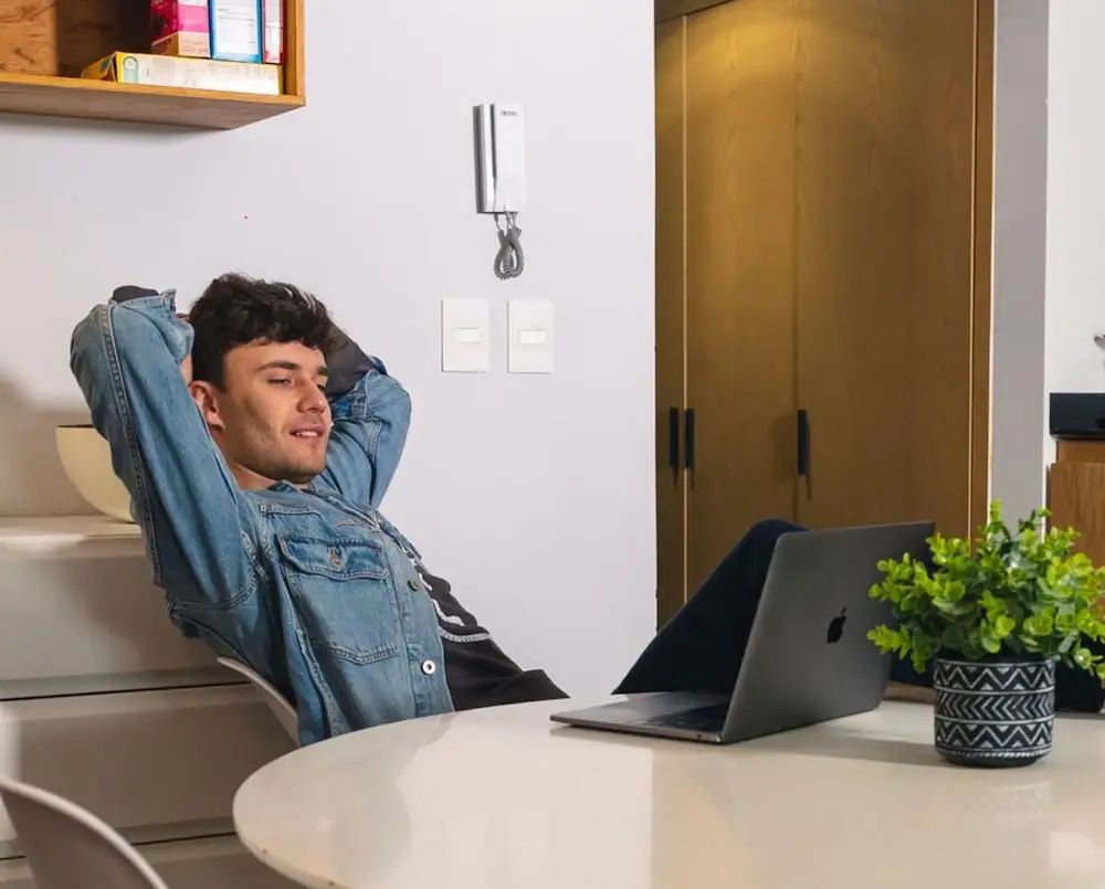 Mann sitzt entspannt vor einem geöffneten Laptop an einem weißen Esstisch.