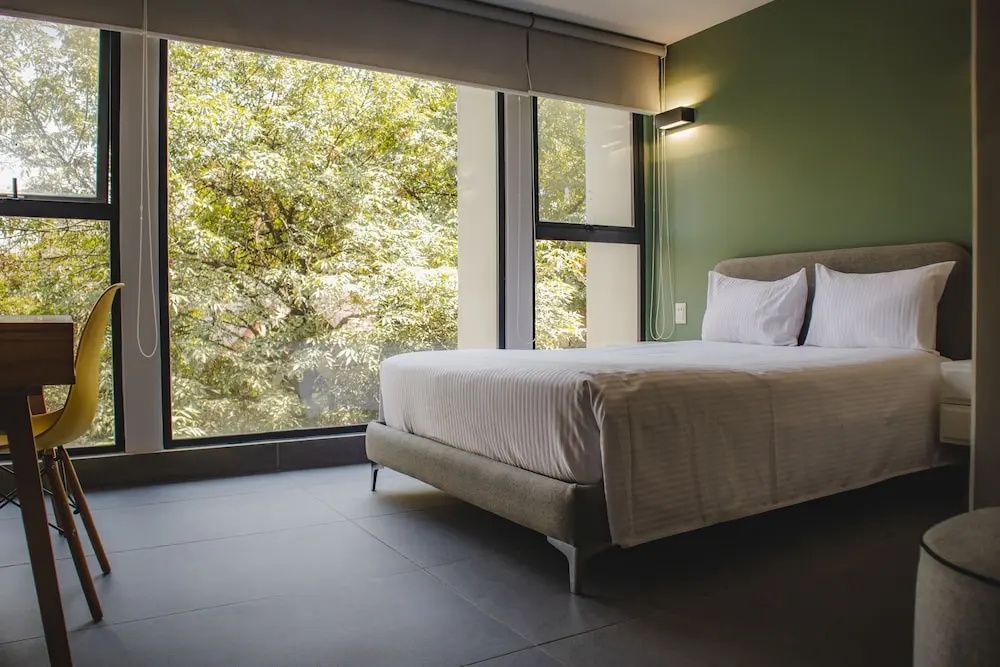 Helles Schlafzimmer mit Doppelbett und Fensterfront mit Blick auf grüne Bäume.