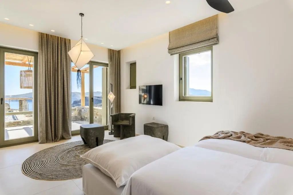 Helles Schlafzimmer mit zwei Betten, Fernseher an der Wand, Sitzmöbeln und Balkontür.