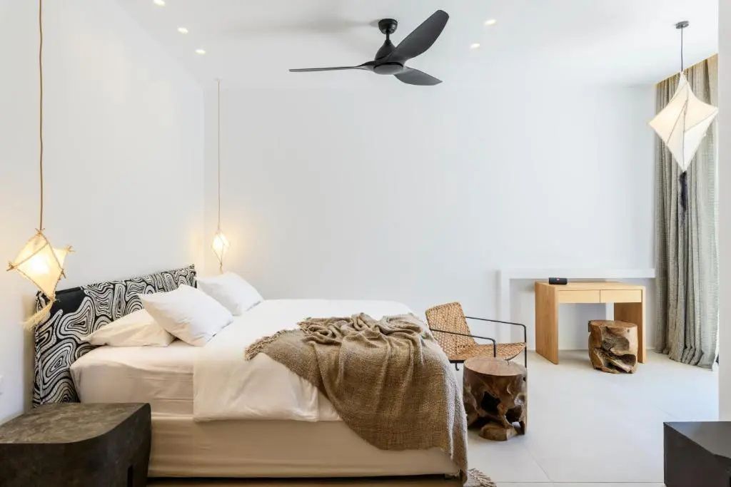 Hell eingerichtetes Schlafzimmer mit Doppelbett, Ventilator an der Decke und Holzstuhl.
