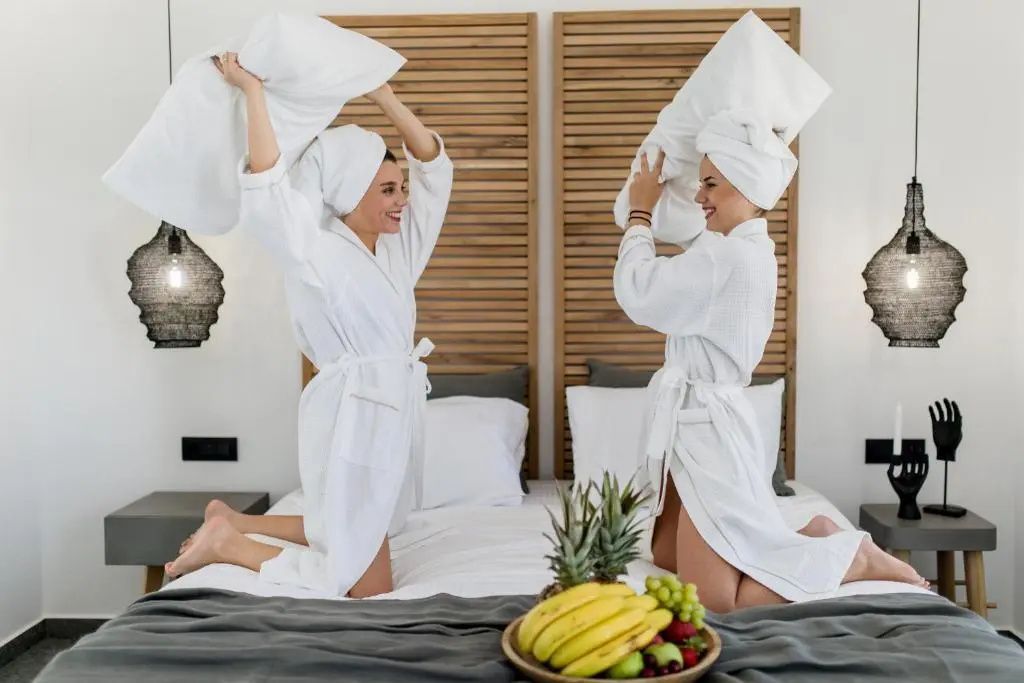 Zwei Frauen in weißen Bademänteln machen eine Kissenschlacht auf einem Hotelbett.