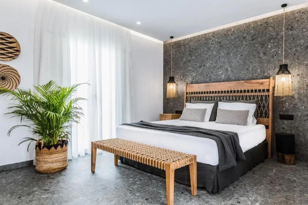 Doppelbett vor einer dunklen Steinwand mit Flechtbank davor und hellen Vorhängen.