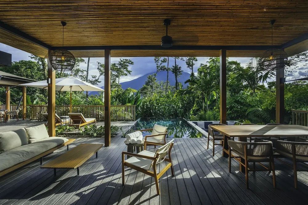 Überdachte Holzterrasse mit Pool, Liegestühlen und Ausblick auf die umgebende Natur und einen Vulkan.