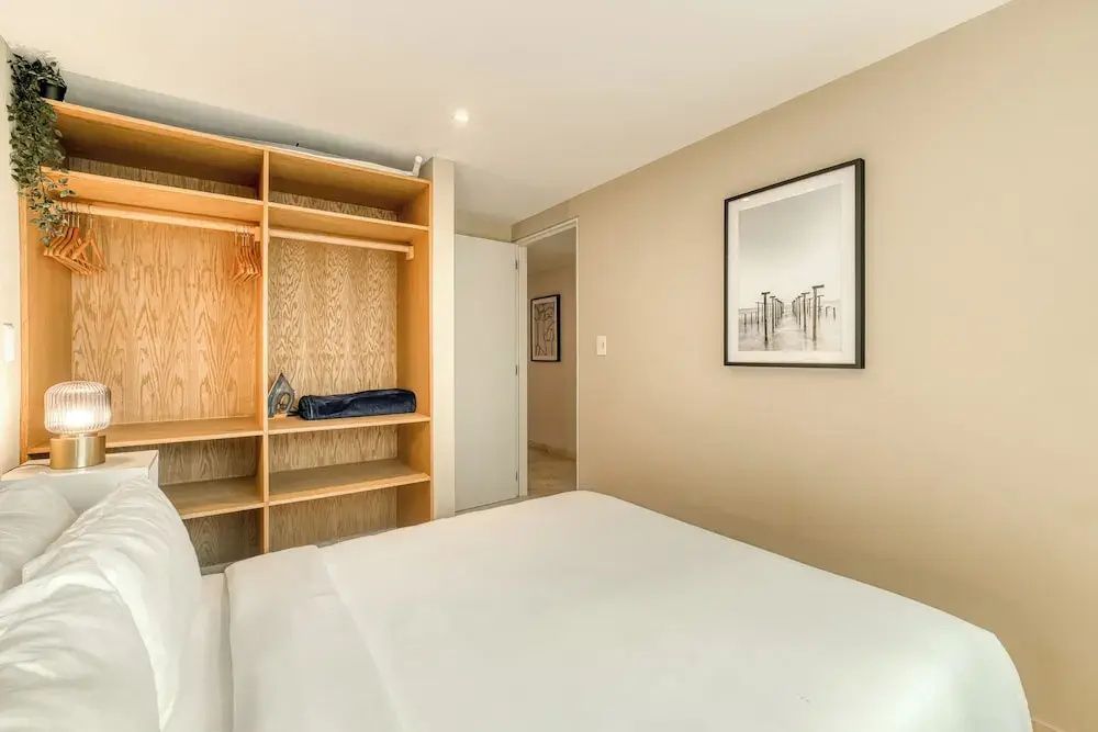 Schlafzimmer mit weißem Bett, offenem Holzkleiderschrank und einem Bild an der beige Wand.