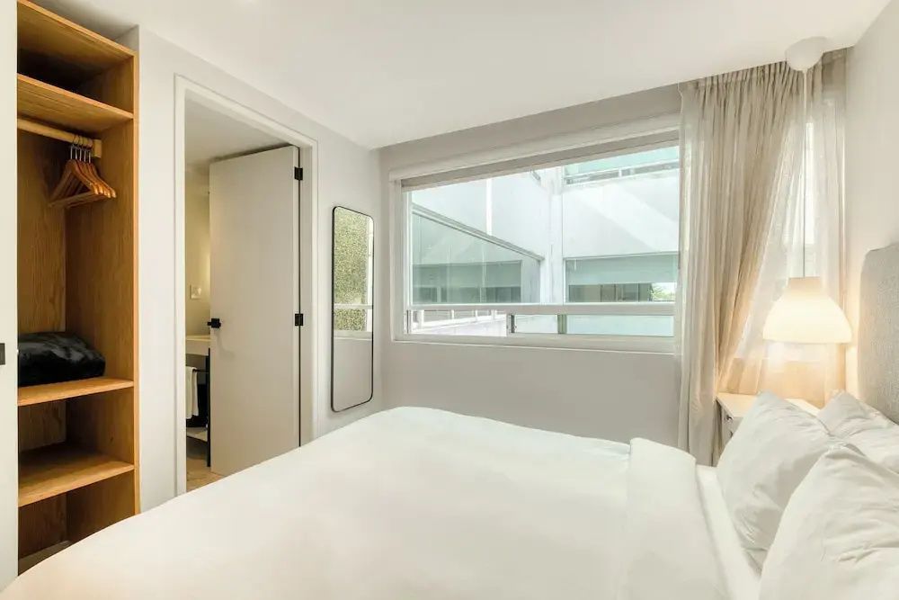 Helles Schlafzimmer mit weißem Bett, einem großen Fenster und einem schmalen Wandspiegel.