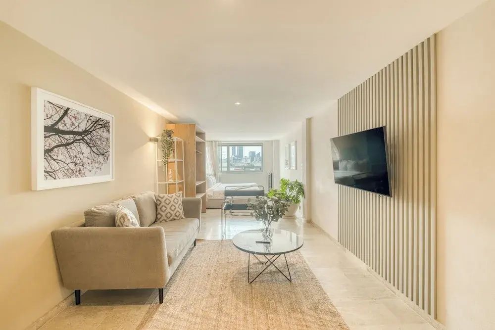 Geräumiges Wohnzimmer mit beigem Sofa, Wand-TV und Blick zum Schlafbereich.