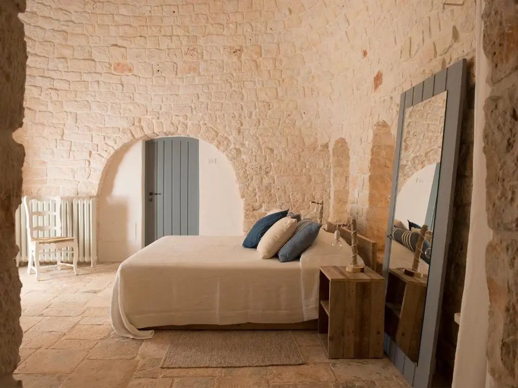 Schlafzimmer mit Natursteinwänden, Doppelbett, Nachttisch und einem großen Standspiegel.