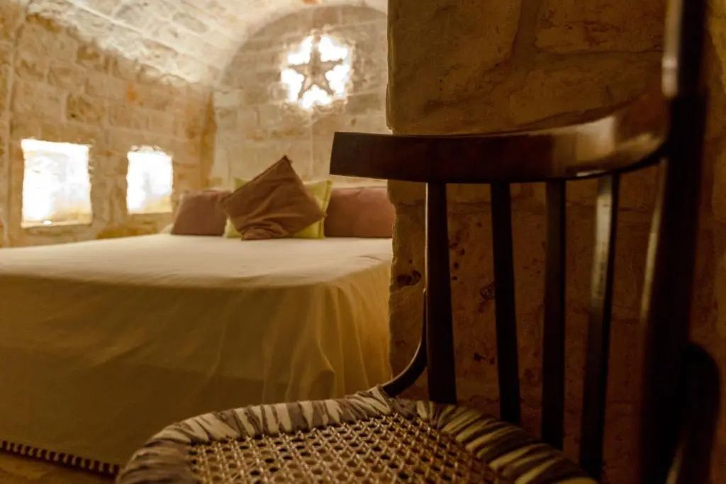 Blick vorbei an einem Holzstuhl auf ein Doppelbett im natursteingefliesten Trullo Zimmer.
