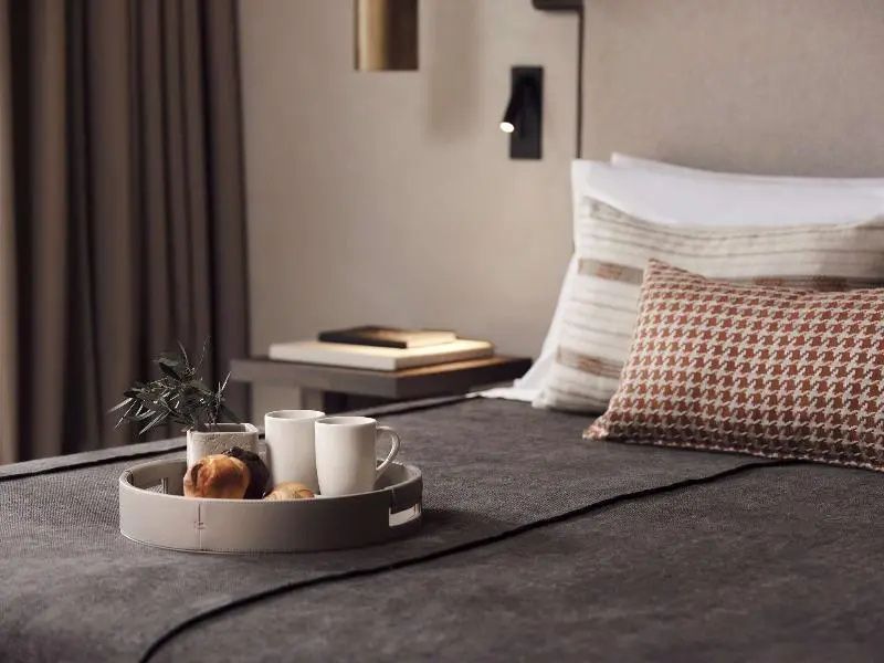 Ein Tablett mit zwei Kaffeetassen und Gebäck auf einem Hotelbett mit Kissen im Hintergrund.