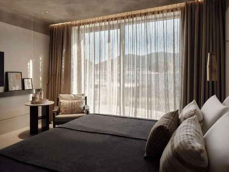 Blick auf das Doppelbett und ein großes Fenster mit transparenten Vorhängen und Sessel.