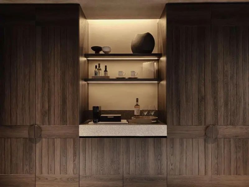 Beleuchtete Nische mit Minibar und Gläsern, eingefasst in eine schlichte Holzverkleidung.
