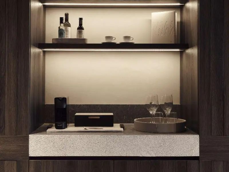 Minibar im Zimmer mit beleuchteten Regalen, Gläsern und Flaschen auf einer Steinplatte.