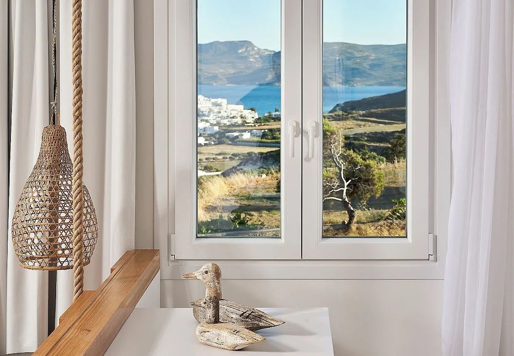 Blick durch ein weißes Fenster auf das Meer, weiße Häuser und eine hügelige Küstenlandschaft.