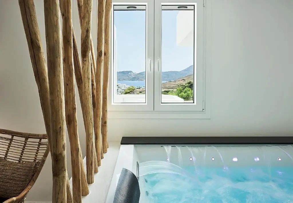 Blick durch ein Fenster auf eine Meeresbucht neben einer befüllten Whirlpool Wanne mit Wasserfall