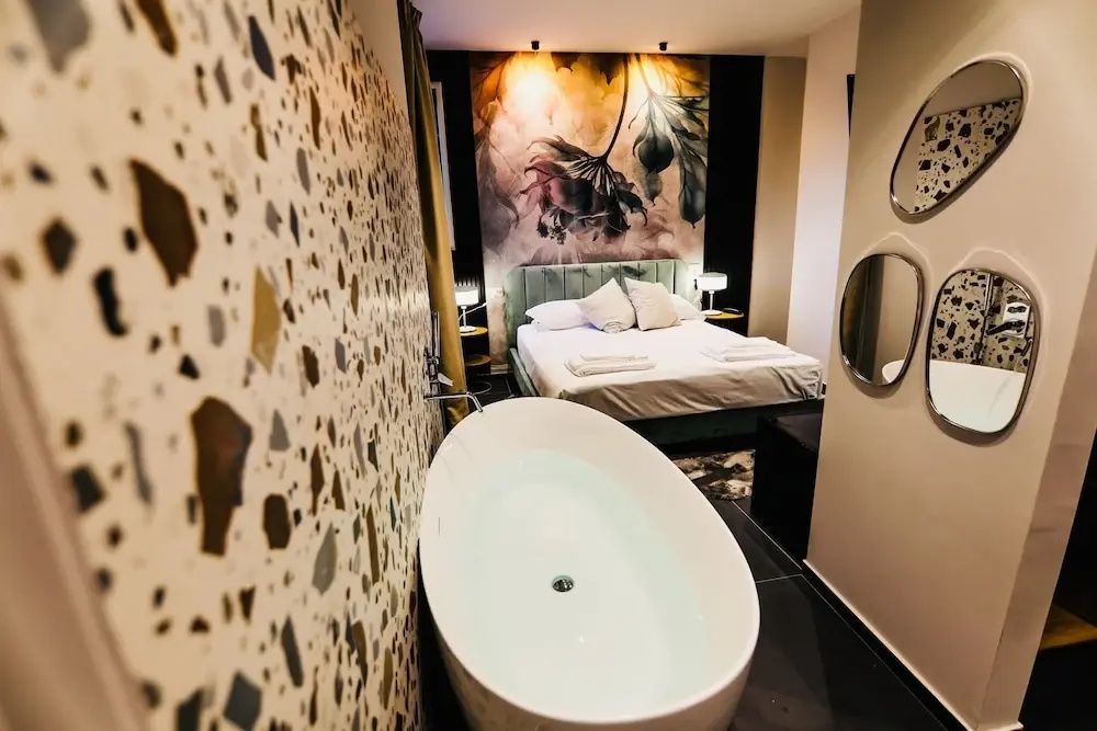 Blick über die gefüllte Badewanne auf das Doppelbett mit Wandbild im Schlafbereich.