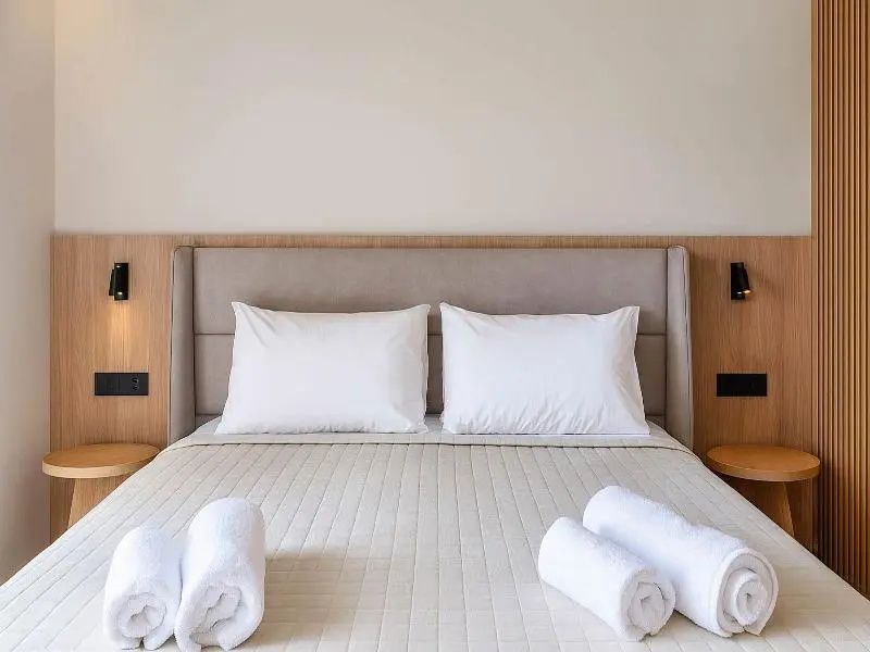 Doppelbett mit gepolstertem Kopfteil, weißen Kissen und ordentlich gerollten Handtüchern.